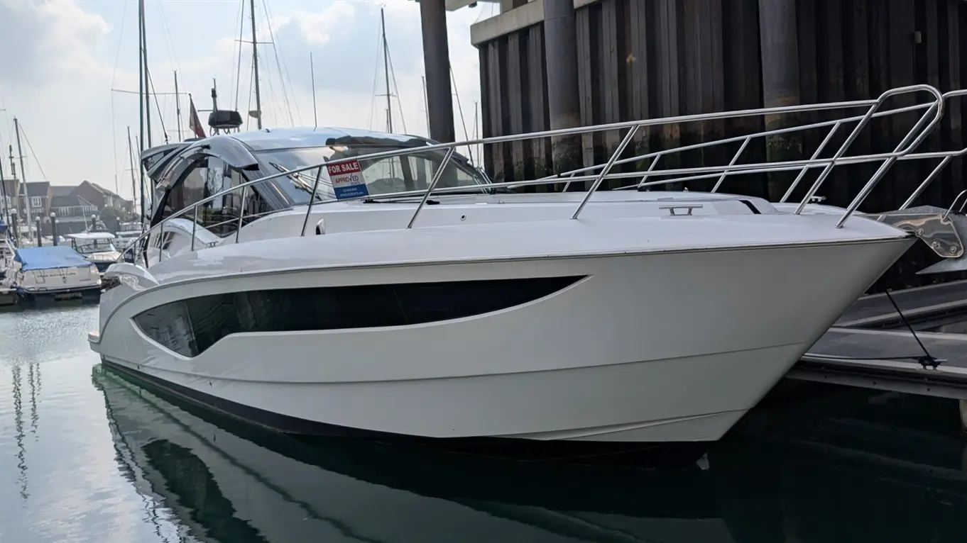 White motorboat docked at a marina, featuring a "For Sale" sign and reflections on the water surface.